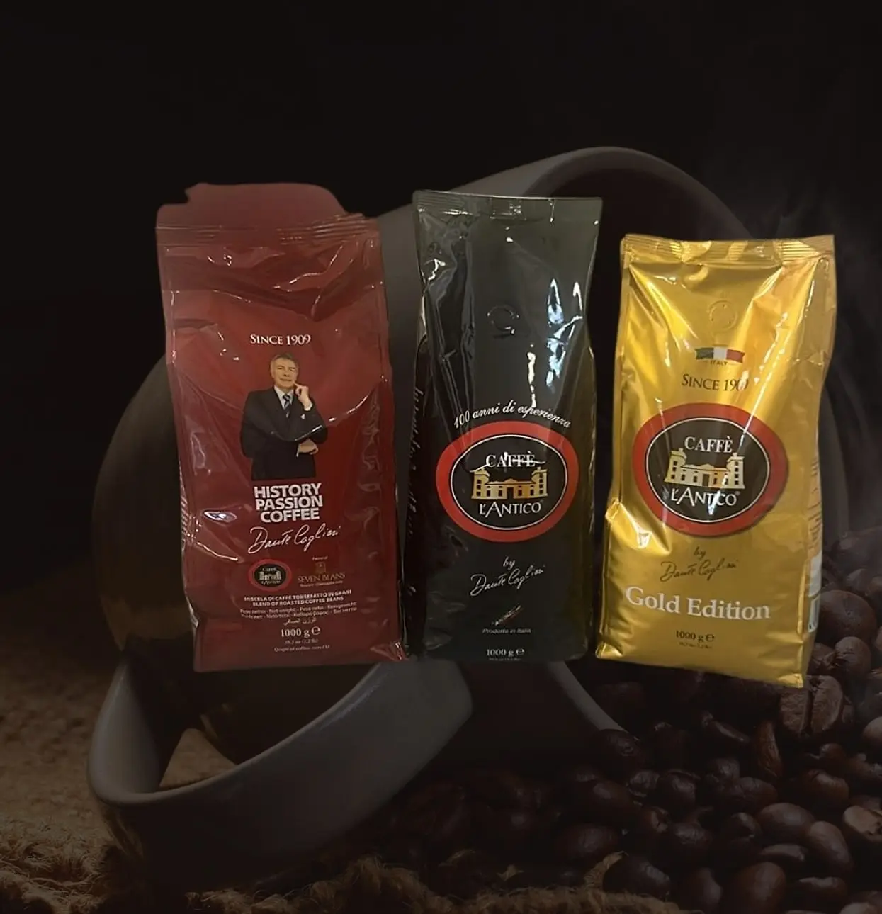 Drei Packungen Caffè L’Antico by Dante Cagliari zu je 1 kg: History Passion Coffee (rot), 100 anni di esperienza (schwarz) und Gold Edition (gold) vor Kaffeebohnen